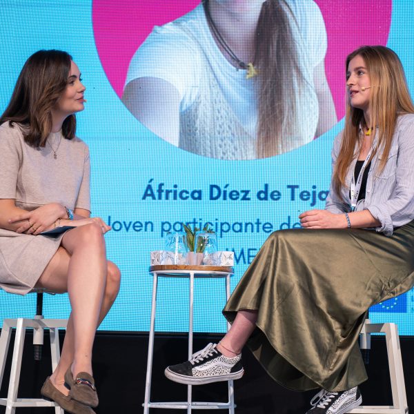 En el escenario, la periodista Eva Baroja entrevista a África Díez, participante del proyecto IMEX. Ambas están sentadas en un taburete.