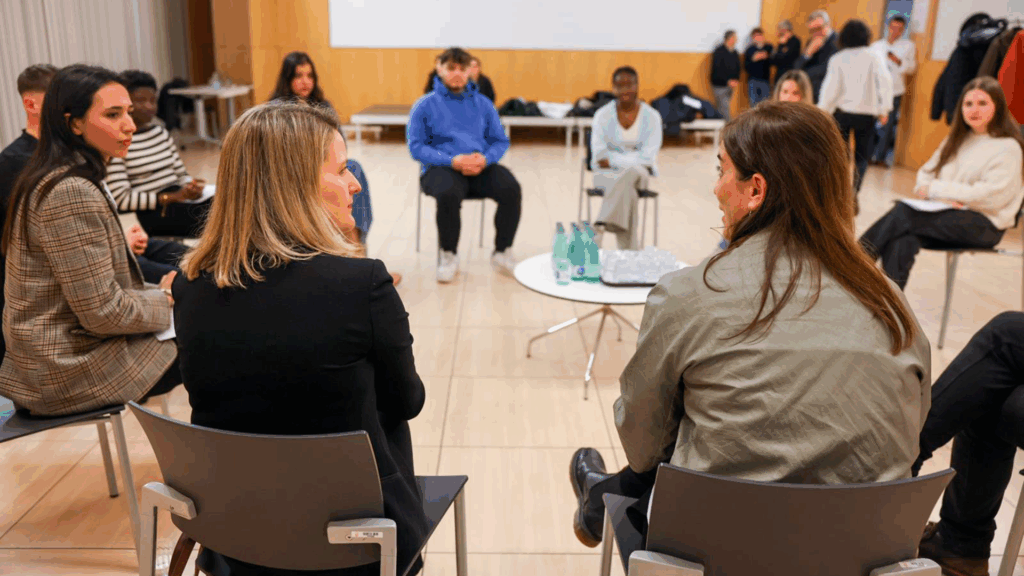Les conselleres de Recerca i Univesitats i de Drets Socials i Inclusió, d'esquenes, assegudes en cercle i conversant amb un grup d'universitaris i universitàries extutelats.