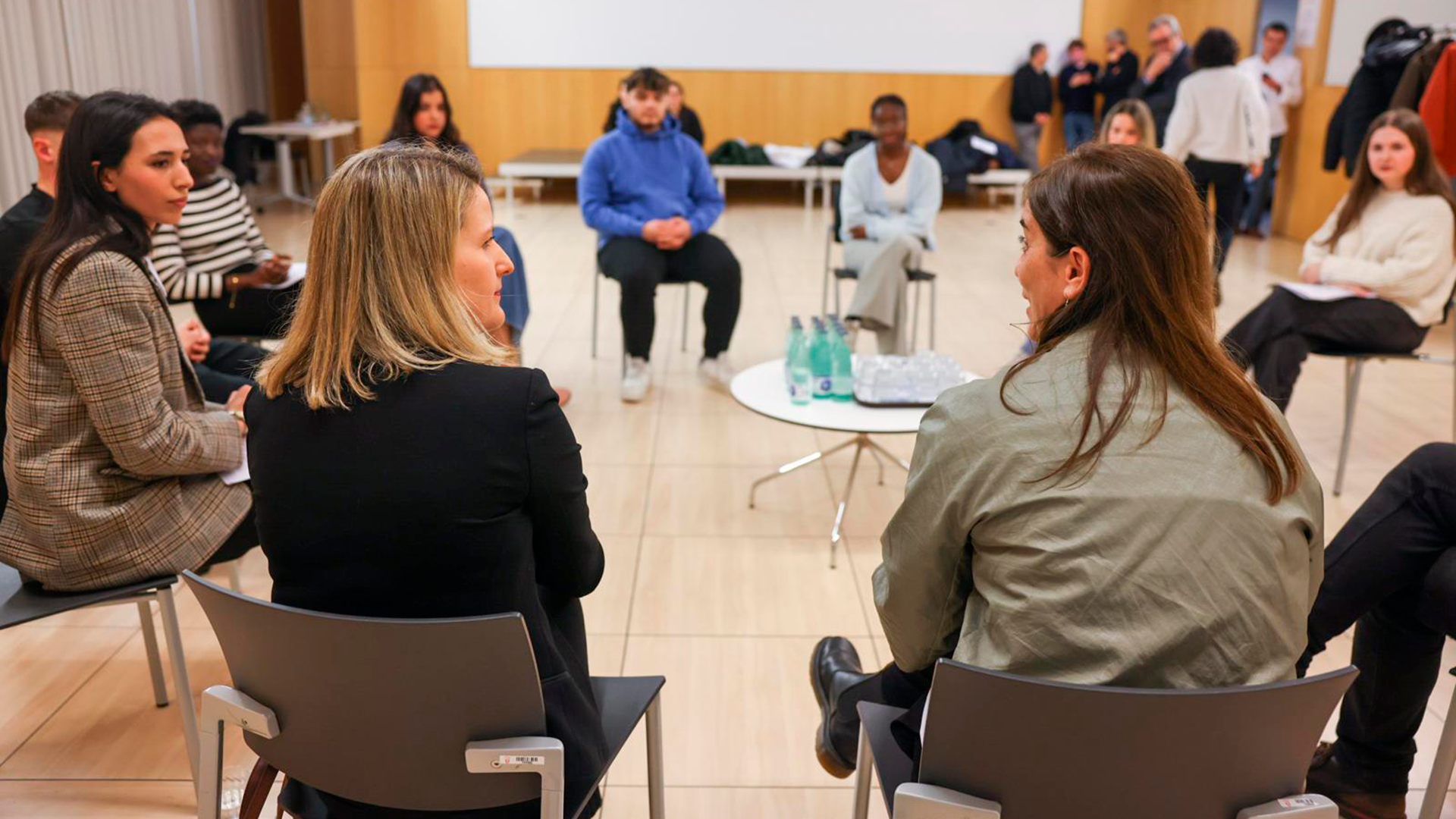 Les conselleres de Recerca i Univesitats i de Drets Socials i Inclusió, d'esquenes, assegudes en cercle i conversant amb un grup d'universitaris i universitàries extutelats.