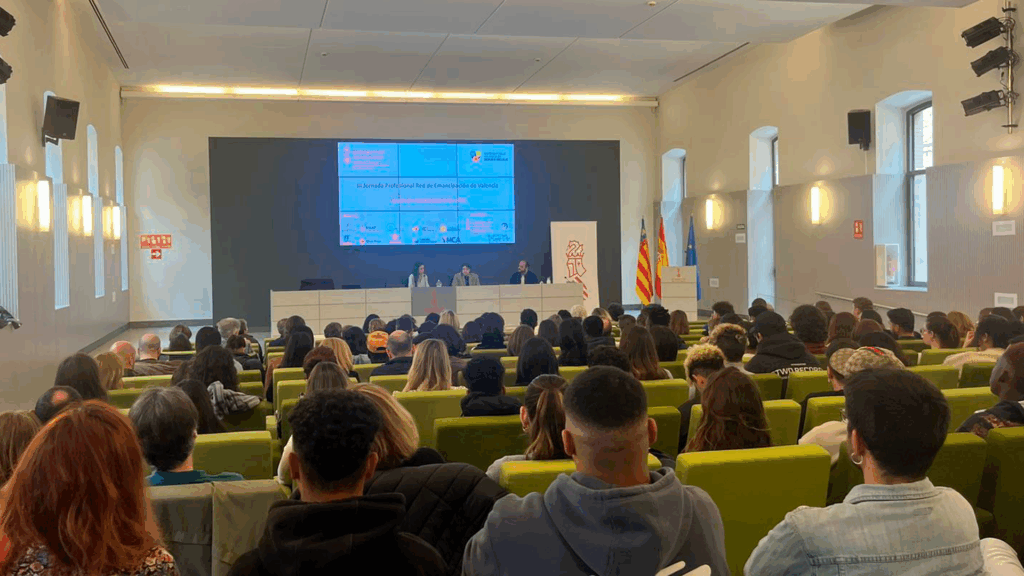 Auditorio de la jornada en la que se ve a los asistentes de espaldas y, a lo lejos, en la mesa del escenario, al equipo dinamizador de Entre Joves.