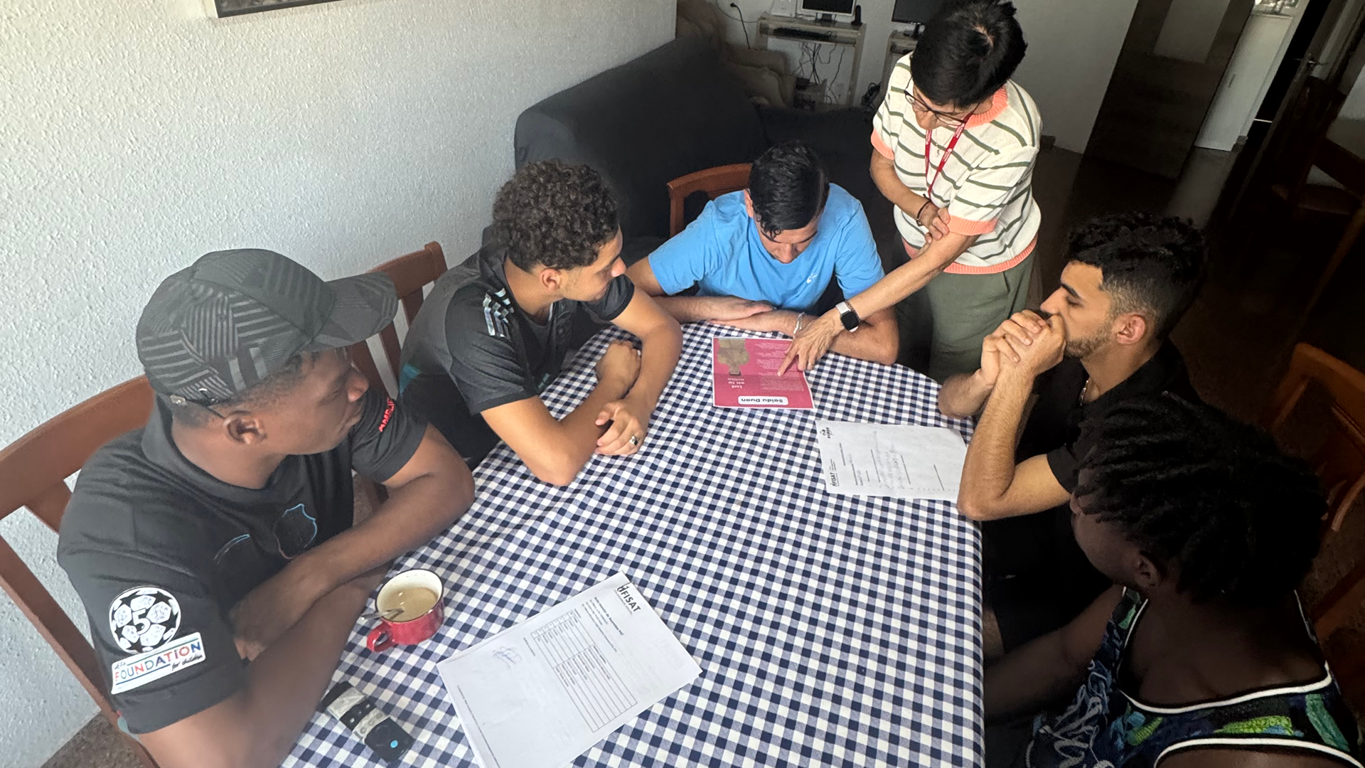 Cinco jóvenes sentados en la mesa de su piso de emancipación atentos al documento de uno de ellos que le entrega una profesional de Fundación Fisat.
