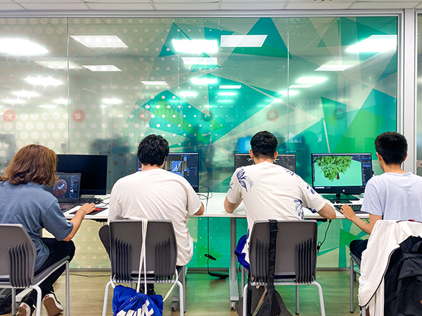 Cuatro jóvenes de espaldas en un aula de informática, cada uno de ellos, con su respectivo ordenador de mesa.