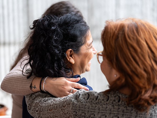 Tres mujeres de espaldas pasándose el brazo por encima del hombro. Están de perfil, pero se aprecia que están sonrientes.
