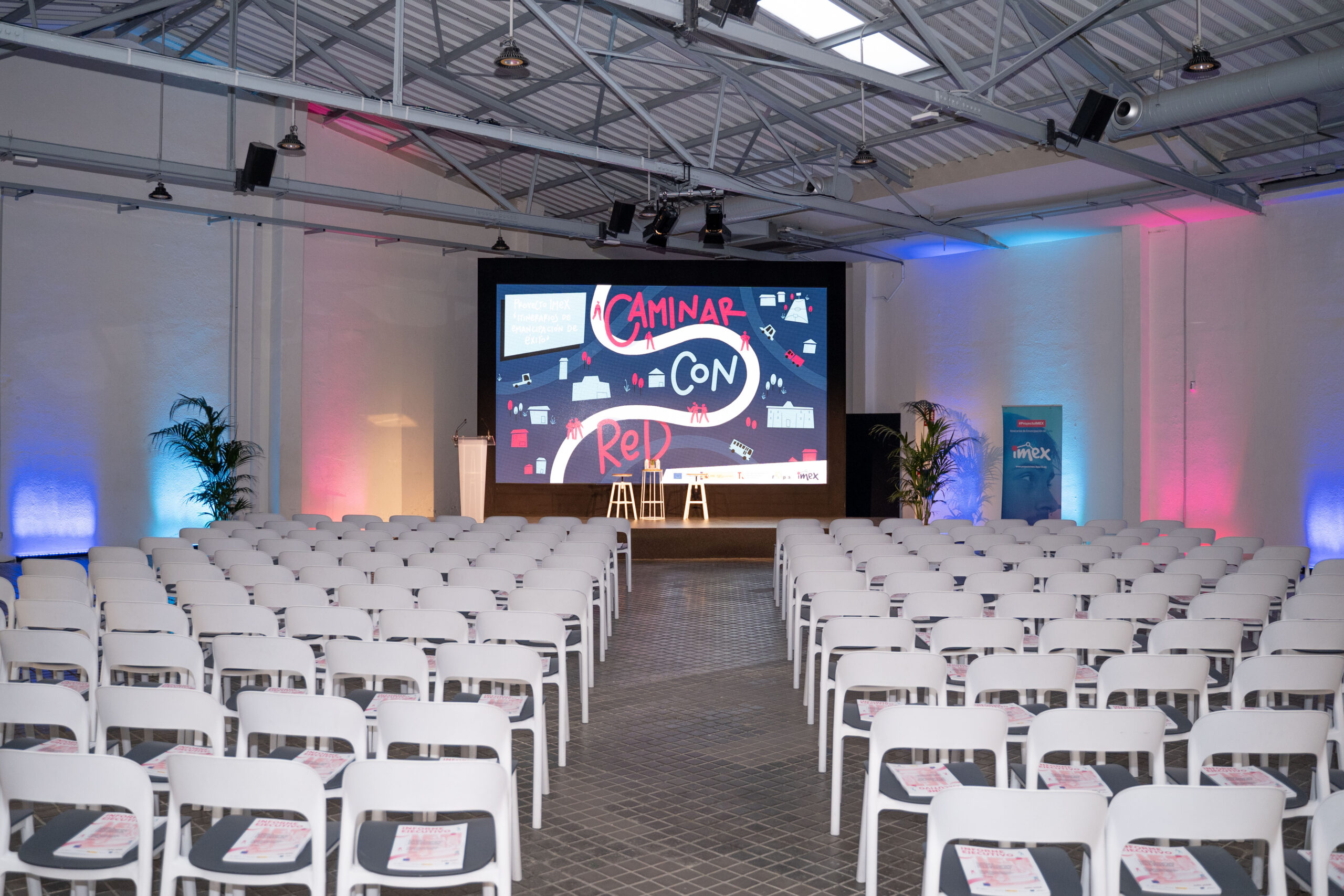 Imagen de la sala del Espacio Rastro en el que se llevaron a cabo las jornadas IMEX de Madrid, vacía. Se ven las sillas y de fondo, la pantalla con el título de las jornadas 'Caminar con red'.