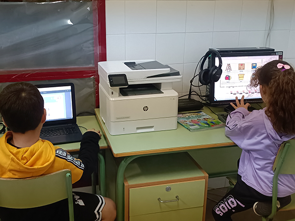 Dos niños, de espaldas, en un aula de informática, cada uno con su ordenador.