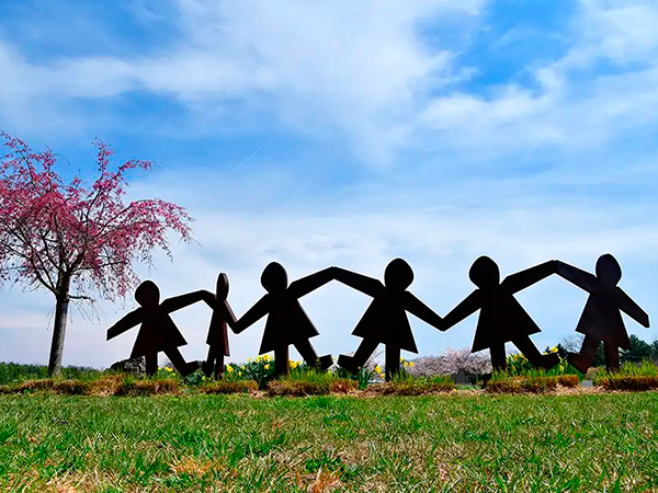 Foto de un parque en el que, entre flores, se aprecian unas figuras de cartón que simulan ser niños o inñas que se dan la mano.