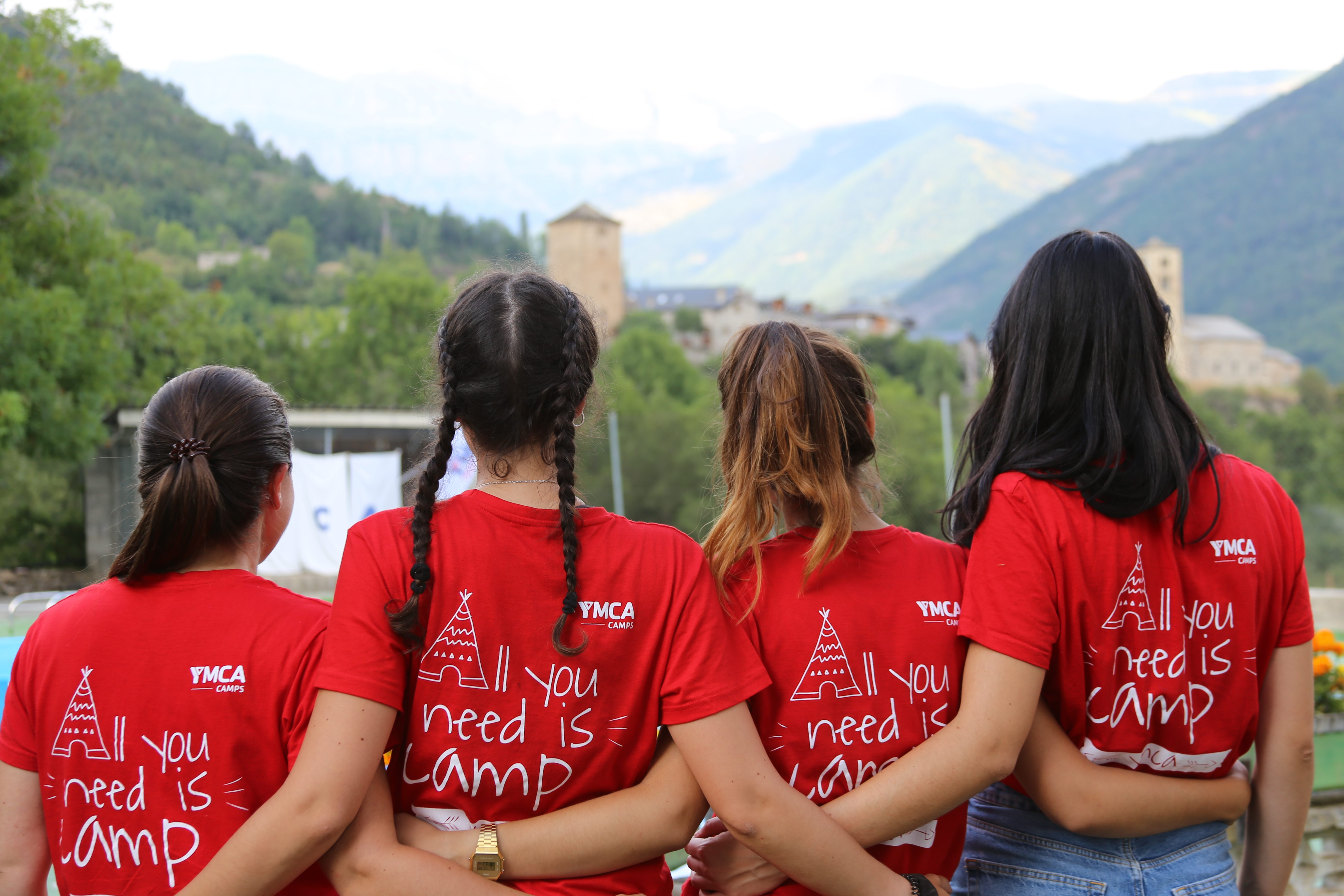 Cuatro chicas de espalda observan un paisaje montañoso. Llevan una camiseta roja de YMCa en la que se lee 'All you need is camp'.