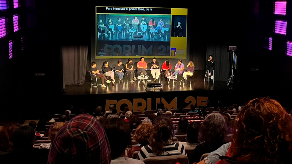 Foto del auditorio desde arriba en la que se ve a los asistentes de espaldas y el escenario de lejos en un momento en el que hay 9 personas en el escenario, además de la intérprete de signos, y tiene el micrófono Aslan Quesada, dinamizador de Entre Joves en FEPA.