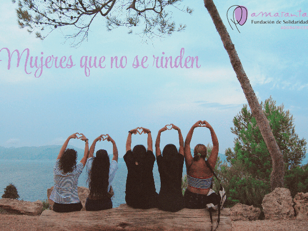 Cinco chicas jóvenes con los brazos en alto y haciendo la forma del corazón con los dedos, sentadas en unas rocas y mirando al mar. En la imagen se lee 'Mujeres que no se rinden' y está incrustado el logo de la Fundación de Solidaridad Amaranta.