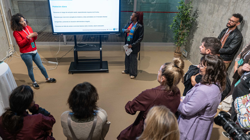 Dos profesionales de entidades exponen una buena práctica, de pie, frente a un grupo de personas, y se acompañan de una pantalla de soporte.
